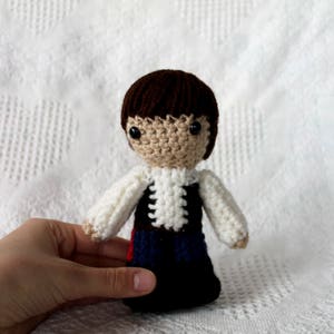 May include: A handmade crochet doll with a brown hair, beige face, and black eyes. The doll is wearing a white shirt with a black vest and blue and red accents. The doll is approximately 5 inches tall.