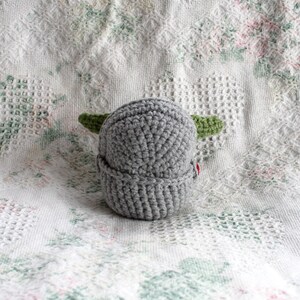 Pu&ograve; includere: Un cappello grigio all'uncinetto con orecchie verdi. Il cappello &egrave; su una coperta bianca all'uncinetto.