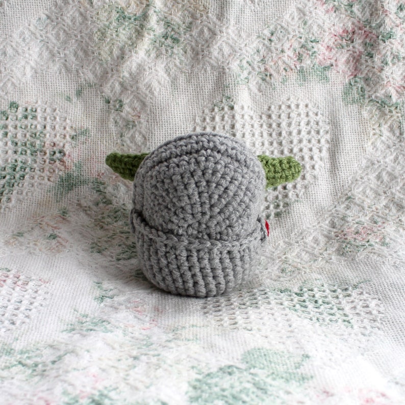 Pu&ograve; includere: Un cappello grigio all'uncinetto con orecchie verdi. Il cappello &egrave; su una coperta bianca all'uncinetto.