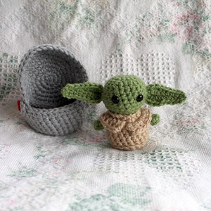 Pu&ograve; includere: Una bambola di Baby Yoda in crochet verde con occhi grandi e vestiti in crochet marroni. La bambola &egrave; seduta su una coperta in crochet bianca accanto a un cesto in crochet grigio.