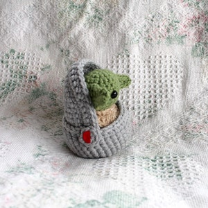 Pu&ograve; includere: Un pupazzo Yoda di uncinetto grigio e verde in una borsa grigia con un bottone rosso. Il pupazzo &egrave; seduto su una coperta di uncinetto bianca.