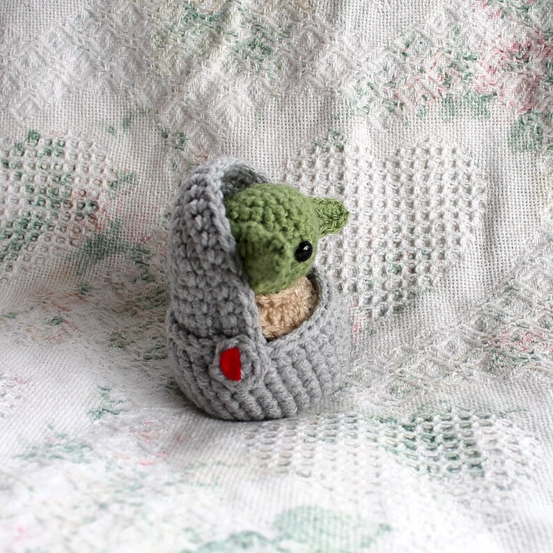 Pu&ograve; includere: Un pupazzo Yoda di uncinetto grigio e verde in una borsa grigia con un bottone rosso. Il pupazzo &egrave; seduto su una coperta di uncinetto bianca.