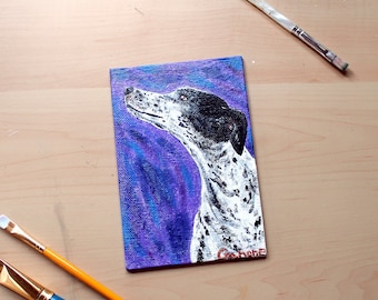 May include: A small rectangular painting of a dog with a black and white spotted coat and a black ear. The background is a blend of purple and blue hues. Two paintbrushes are visible, one with a white handle and the other with a blue handle.