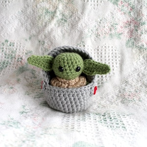 Pu&ograve; includere: Un giocattolo di peluche all'uncinetto di un bambino alieno verde in un baccello grigio. Il giocattolo ha occhi grandi ed &egrave; seduto su una coperta bianca all'uncinetto.