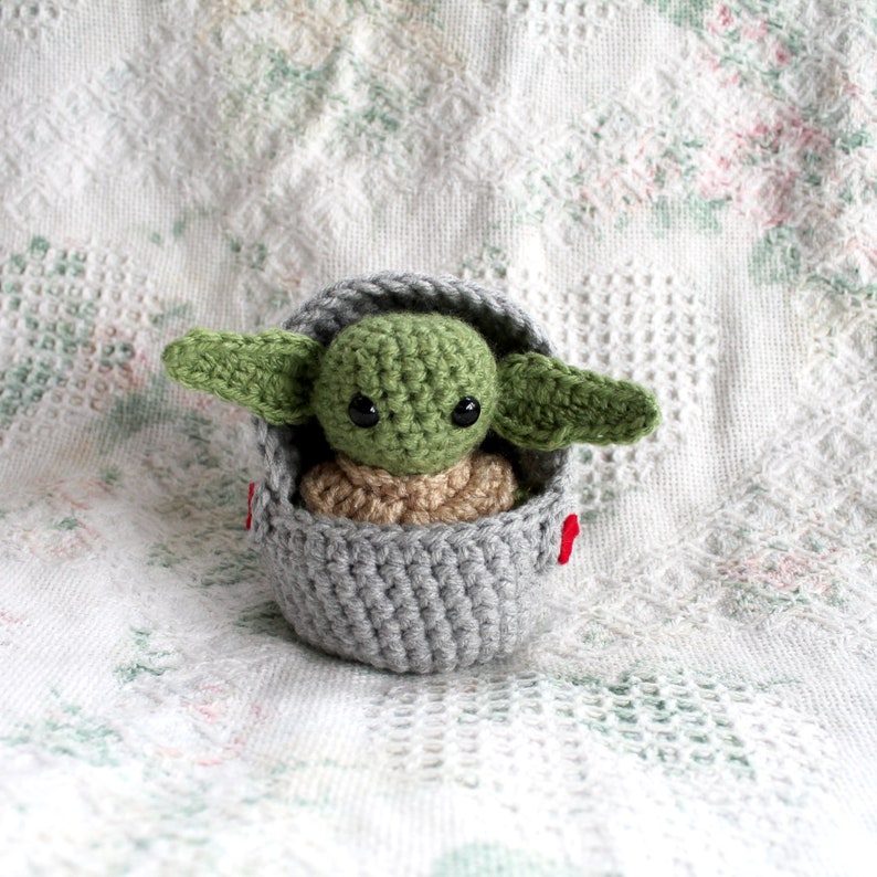 Pu&ograve; includere: Un giocattolo di peluche all'uncinetto di un bambino alieno verde in un baccello grigio. Il giocattolo ha occhi grandi ed &egrave; seduto su una coperta bianca all'uncinetto.