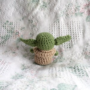 Pu&ograve; includere: Un giocattolo di Baby Yoda in crochet verde e marrone. Il giocattolo &egrave; seduto su una coperta di crochet bianca.