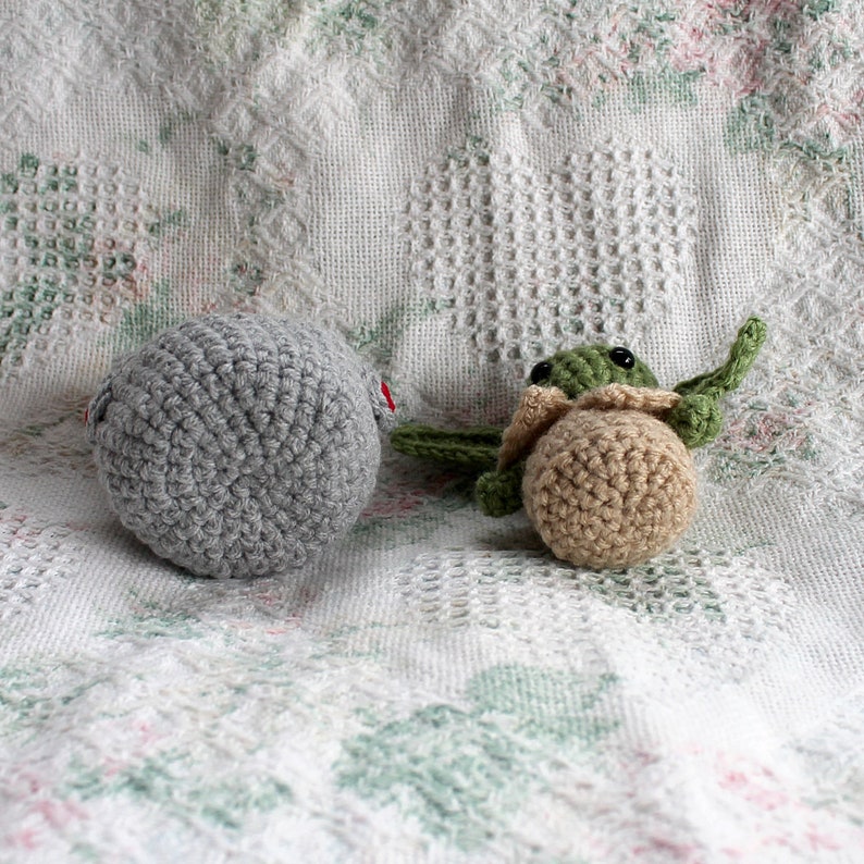Pu&ograve; includere: Due giocattoli di peluche all'uncinetto su una coperta bianca all'uncinetto. Uno &egrave; una palla grigia e l'altro &egrave; un Yoda verde e marrone.