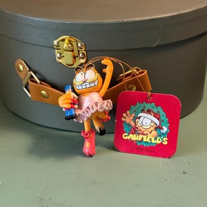 Puede incluir: Un adorno navideño de Garfield. El gato naranja viste un tutú rosa y zapatillas de ballet, sosteniendo una botella azul. Una etiqueta roja con el logo de Garfield y el texto "© 1996 Paws" está adjunta.