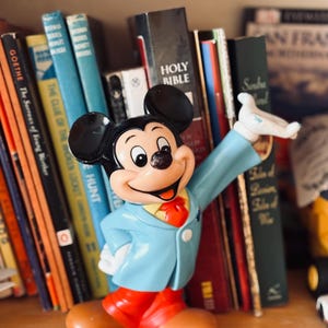 May include: A vintage Mickey Mouse figurine, wearing a light blue jacket, red pants, and brown shoes, stands on a shelf in front of a collection of books. The figurine has black ears and a smiling expression, with one arm raised.