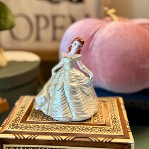 Puede incluir: Figura de porcelana de una mujer con un vestido blanco roto y detalles dorados. La figura tiene el pelo rojo y posa con los brazos ligeramente extendidos. Se encuentra sobre una caja de madera decorativa con intrincados diseños incrustados.