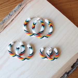 Op de afbeelding: Drie paar kralen oorringen in verschillende maten. De oorringen zijn wit met zwarte, rode, gele en turquoise accenten. De kralen zijn gerangschikt in een chevron-patroon.
