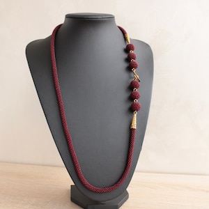 Peut inclure: Un long collier de perles bordeaux avec un fermoir doré et cinq perles dorées. Le collier est présenté sur un mannequin noir.