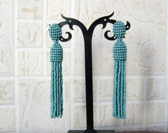 May include: A pair of teal blue beaded tassel earrings displayed on a black earring stand. The earrings are long and dangle.