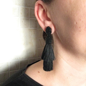 May include: A pair of black tassel earrings with a beaded top. The earrings are dangling from the earlobe of a person.