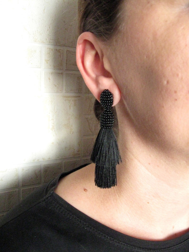 May include: A pair of black tassel earrings with a beaded top. The earrings are dangling from the earlobe of a person.