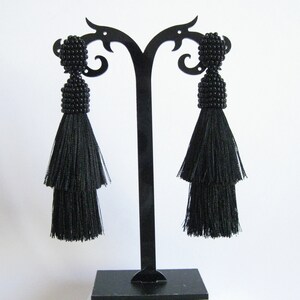 May include: A pair of black tassel earrings with beaded tops. The earrings are displayed on a black earring stand.