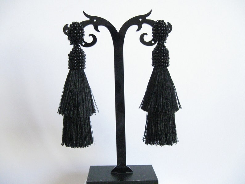 May include: A pair of black tassel earrings with beaded tops. The earrings are displayed on a black earring stand.