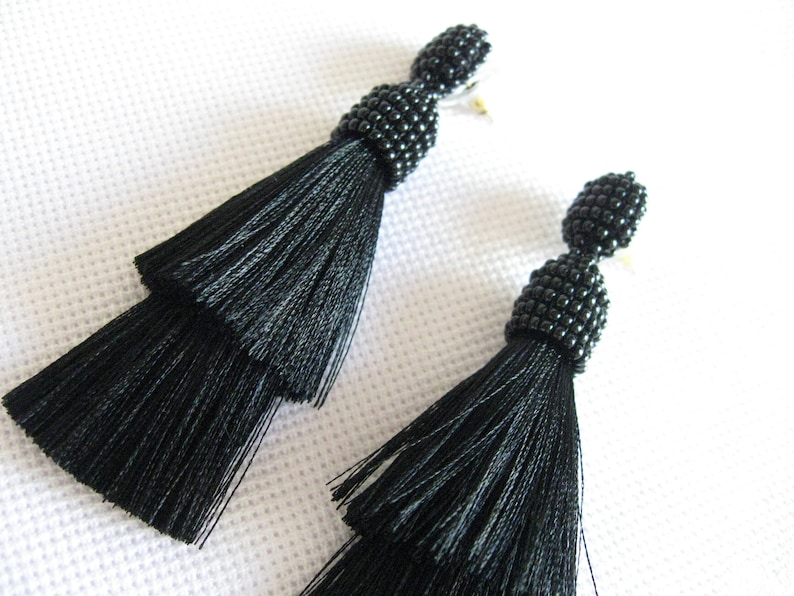 May include: A pair of black tassel earrings with black bead accents. The earrings are made of silk and have a layered tassel design.