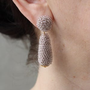 May include: A pair of dangle earrings with a round bead at the top and a teardrop-shaped bead below. The beads are made of small, white beads and have a light pink hue.