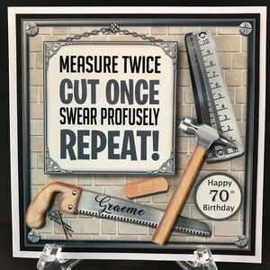 Op de afbeelding: Een vierkante wenskaart met een bordje met de tekst MEASURE TWICE CUT ONCE SWEAR PROFUSELY REPEAT! De kaart bevat illustraties van een metalen liniaal, een hamer, een zaag met het opschrift Graeme, een pleister en Happy 70th Birthday.