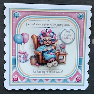Op de afbeelding: Een verjaardagskaart met een cartoonillustratie van een vrouw in een blauwe badjas en krulspelden, zittend op een stoel met een kopje thee. De kaart heeft een roze en blauwe achtergrond met witte stippen en de tekst "Ik had niet van plan om vandaag iets te doen... Ontspan, het is je verjaardag! Tot nu toe loopt alles volgens schema!"