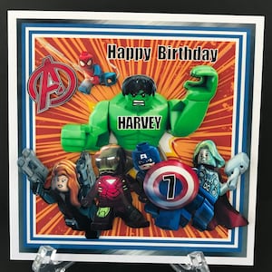 May include: A birthday card featuring a Lego Hulk with the name Harvey on his chest. The Hulk is surrounded by other Lego superheroes including Spiderman, Iron Man, Captain America, Black Widow, and Thor. The card has a red, yellow, and blue comic book background with the words "Happy Birthday" at the top.