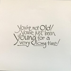 Op de afbeelding: Een vierkante witte kaart met zwarte handgeschreven tekst die luidt: You're not Old! You've just been Young for a very long time! De belettering gebruikt een mix van cursieve en gedrukte stijlen met decoratieve onderstrepingen onder de woorden very en long.