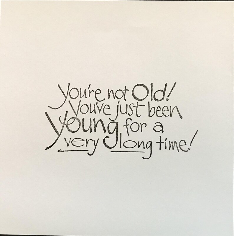 Op de afbeelding: Een vierkante witte kaart met zwarte handgeschreven tekst die luidt: You're not Old! You've just been Young for a very long time! De belettering gebruikt een mix van cursieve en gedrukte stijlen met decoratieve onderstrepingen onder de woorden very en long.