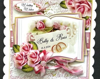 Op de afbeelding: Een witte trouwkaart met een roze en witte kant achtergrond. De kaart heeft een boek met de woorden "Wedding Wishes" en "Sally & Ross" met de datum "12.8.2024". De kaart is versierd met roze rozen en een roze lint.