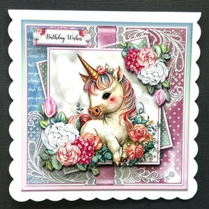 Op de afbeelding: Een handgemaakte verjaardagskaart met een wit eenhoorn met een roze en gouden manen en hoorn, omringd door roze, witte en rode bloemen. De kaart heeft een gegolfde rand en de woorden "Birthday Wishes" staan ​​gedrukt op de voorkant.
