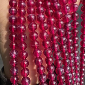 May include: Close-up of strands of deep red gemstone beads. Each bead features a star-like pattern, creating a unique visual effect. The beads are round and strung together, possibly for jewellery making or crafting. The beads are translucent.