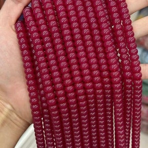 Könnte beinhalten: Eine Strähne aus tief roten Glasperlen, jede Perle ist rund und leicht durchscheinend. Die Perlen sind zu einer langen, durchgehenden Strähne aufgereiht.