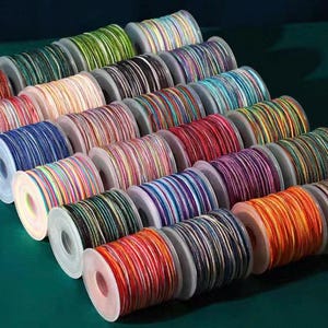 Op de afbeelding: Een verzameling spoelen met kleurrijk koord, elk strak gewikkeld. De koorden tonen een verscheidenheid aan kleuren, waaronder rood, blauw, groen en geel, waardoor een regenboogeffect ontstaat. De spoelen zijn in nette rijen op een donkergroen oppervlak gerangschikt.