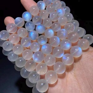 May include: A collection of round, translucent moonstone beads. Each bead has a soft, pearly sheen with flashes of blue light. The beads are held in a hand, showcasing their size and color variations.