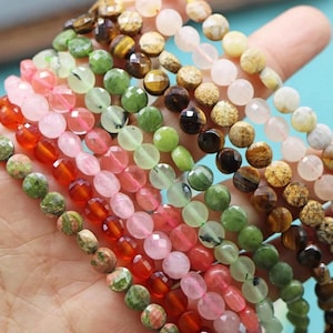May include: Assortment of round gemstone beads in various colors, including green, pink, red, brown, and white. The beads are strung on a string and are displayed on a hand.
