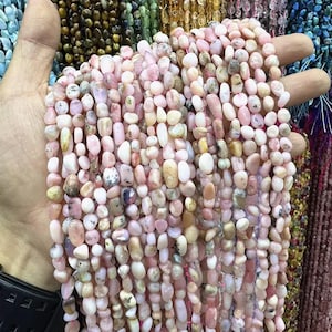 Peut inclure: Plusieurs brins de perles en opale rose de différentes formes et tailles. Les perles varient du rose pâle au blanc crème, certaines présentant des motifs mouchetés. Les perles sont tenues dans une main, avec d'autres brins de perles en arrière-plan.