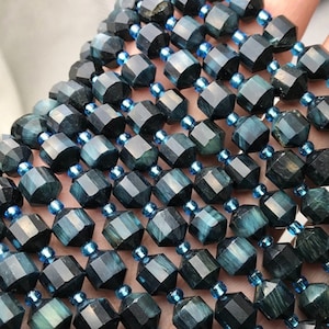 May include: Close-up of multiple strands of faceted, dark teal gemstone beads. Each bead is a geometric shape with a dark teal color and is separated by small, light blue beads. The beads are strung together, creating a textured surface.