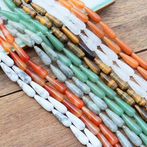 May include: Rows of teardrop-shaped gemstone beads in various colors, including orange, white, green, gray, and tiger's eye. The beads are arranged on a wooden surface, showcasing their polished surfaces and natural variations. Ideal for jewelry making.
