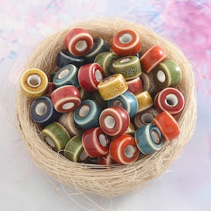 Pode incluir: Uma coleção de contas de cerâmica coloridas num ninho tecido. As contas são cilíndricas com um orifício central e vêm em várias cores, incluindo vermelho, azul, verde e amarelo. As contas são provavelmente usadas para fazer joias ou artesanato.