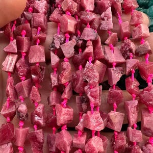 Puede incluir: Primer plano de hilos de cuentas de piedras preciosas de rodocrosita rosa. Las cuentas están talladas de forma tosca y ensartadas en un cordón rosa brillante. Las piedras muestran formas naturales irregulares y diferentes tonos de rosa y rojo. Ideal para la creación de joyas.