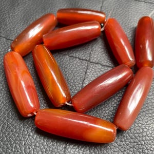 Può includere: Una collana di perline allungate e lucide nei toni del rosso e dell'arancione. Le perline hanno una finitura liscia e lucida e sono infilate insieme con un filo bianco. Le perline sono di forma cilindrica e sembrano essere fatte di un materiale traslucido.