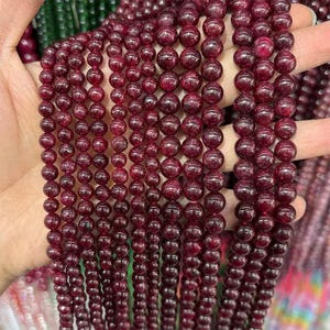 Peut inclure: Gros plan de plusieurs brins de perles de pierres précieuses rondes, rouge foncé. Les perles, de tailles variées, sont enfilées pour un aspect texturé. Elles sont tenues dans une main, mettant en valeur leur couleur et leur éclat. Idéal pour la création de bijoux.