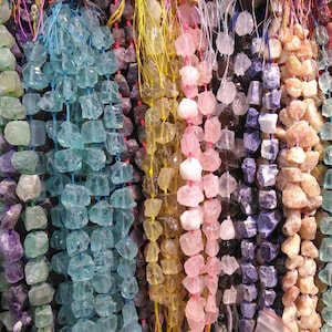 Peut inclure: Plusieurs rangées de pierres précieuses de différentes couleurs, dont le bleu, le jaune, le rose, le violet et le beige. Chaque rangée est composée de pierres brutes de forme irrégulière, enfilées sur des cordons colorés. Les pierres sont exposées de près.