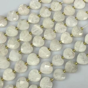 May include: Strands of heart-shaped, off-white gemstone beads. Each bead is faceted and connected with gold-colored metal findings. The beads are strung together, creating a repeating pattern of hearts.