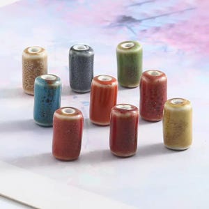 Könnte beinhalten: Zehn Keramikperlen in verschiedenen Farben, darunter Blau, Rot, Grün und Beige. Jede zylindrische Perle hat einen weißen Rand und ein Loch in der Mitte. Die Perlen haben eine glänzende Oberfläche und sind auf einer weißen Oberfläche angeordnet. Maße in Zentimetern.