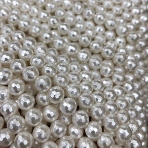 May include: A close-up of a large quantity of white, round, faceted beads. The beads have a pearlescent finish and are arranged in a random pattern.