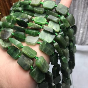 Puede incluir: Una hebra de cuentas de jade verde oscuro, cada cuenta es rectangular y pulida. Las cuentas están enhebradas en un cordón verde.