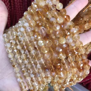 May include: Strands of faceted citrine gemstone beads in shades of yellow and amber. The beads are round and have a sparkling, polished appearance. The gemstones are held in a hand, showcasing their color and texture. Ideal for jewelry making.