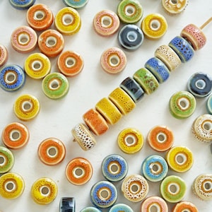 May include: A selection of brightly coloured ceramic donut-shaped beads in shades of yellow, orange, blue, green, and pink. Some beads are strung on a wooden skewer, others are loose. These beads are suitable for jewellery making.