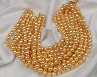 May include: A collection of golden-colored, round beads, likely pearls, arranged in multiple strands. The beads are uniform in size and have a lustrous sheen. The strands are resting on a white, textured fabric, creating a contrast in color and texture.